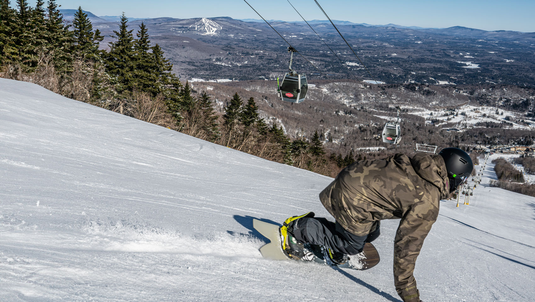 Snowboarder carving a turn on a mountain ski slope with gondolas and a distant ski resort