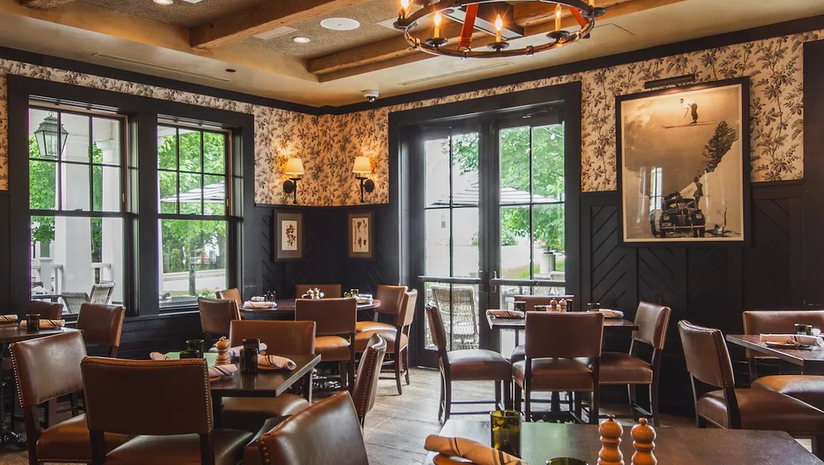 Warm, upscale restaurant dining room with dark wood paneling, leather chairs, floral wallpaper, and set tables beside large windows overlooking outdoor greenery.
